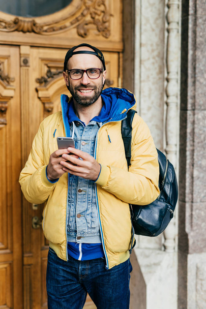 Young traveler in trendy clothes standing in museum having happy look to be there. Cheerful bearded male in yellow jacket holding rucksack on his back holding mobile visiting regional museum - Photo, Image