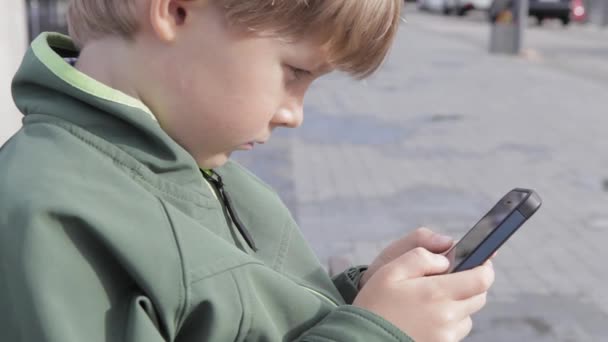Młody chłopak grać na smartfonie w mieście. Chłopiec z telefonu komórkowego. Mały chłopiec na ulicy gry z smartphone - Materiał filmowy, wideo