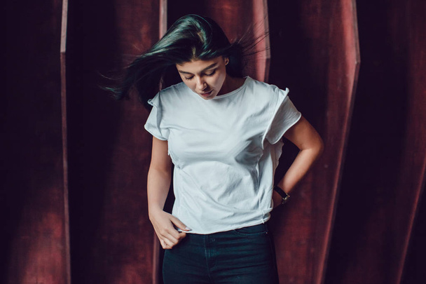 Attractive brunette woman in a white t-shirt stands on a dark wooden wall background. Mock-up. - Фото, зображення