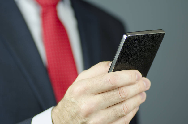 Una vista de cerca de la mano de un gerente en un traje y con una corbata roja usando un teléfono inteligente negro
 - Foto, imagen
