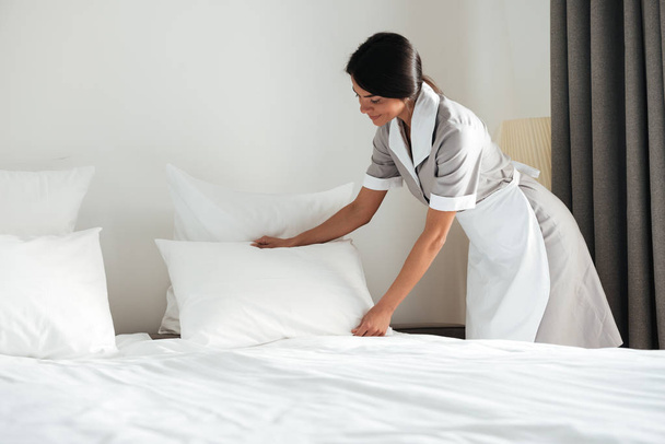 Young hotel maid setting up pillow on bed - Фото, зображення