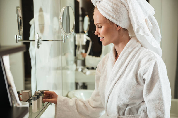 Woman in bathrobe looking at cosmetics near mirror - Фото, зображення