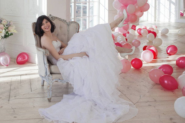 Young woman in wedding dress in luxury interior with a mass of pink and white balloons, sitting in the chair. Charming young bride brunette with short haircut in stylish Quinceanera dresses. - Φωτογραφία, εικόνα