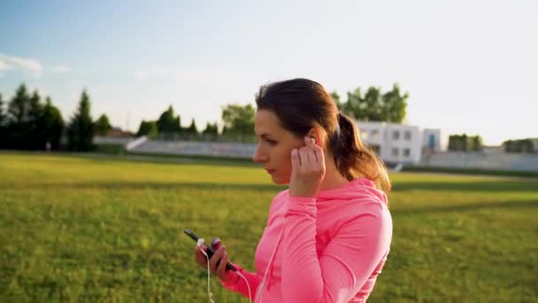 Woman inserts headphones into ears, preparing for a run at the stadium - Séquence, vidéo
