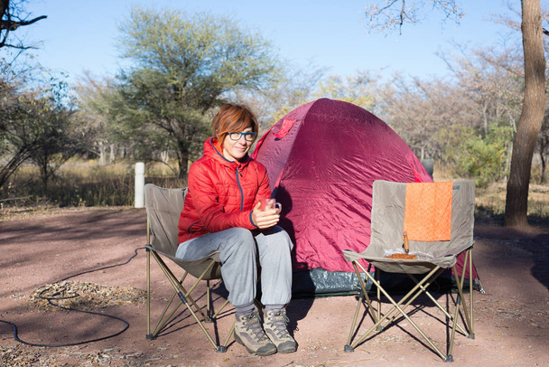 Woman drinking hot coffee mug while relaxing in camping site. Tent, chairs and camping gears. Outdoor activities in summer. Adventure traveling in South African National Park. - Photo, Image