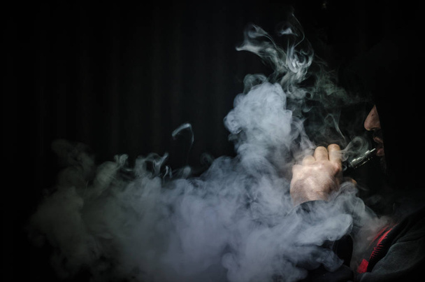 Vaping man holding a mod. A cloud of vapor. Black background. Vaping an electronic cigarette with a lot of smoke - Photo, Image