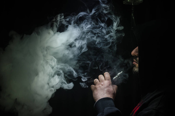 Hombre aspirante sosteniendo un mod. Una nube de vapor. Fondo negro. Vapeando un cigarrillo electrónico con mucho humo
 - Foto, Imagen