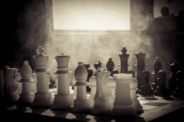 Концепция шахматной настольной игры бизнес-идей, конкуренции и стратегических идей. Шахматные фигуры на темном фоне с дымом и туманом. Выборочный фокус. Рука кладет фигуру на борт
 - Фото, изображение