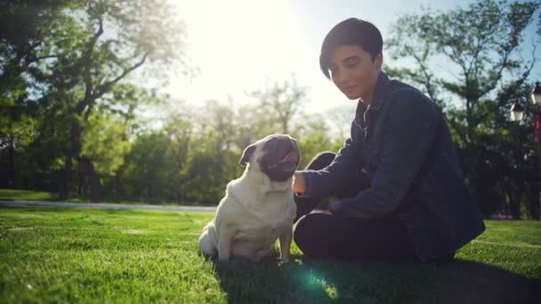 Young stylish girl caress happy dog of pug breed park summer sun slow motion - Záběry, video
