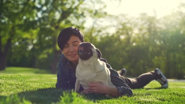 Young beautiful girl lies grass park caress happy puppy pug summer sun - Záběry, video