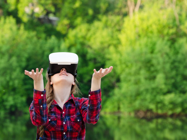 Meisje in de helm van virtual reality tegen de achtergrond van de natuur. Handen omhoog - Foto, afbeelding