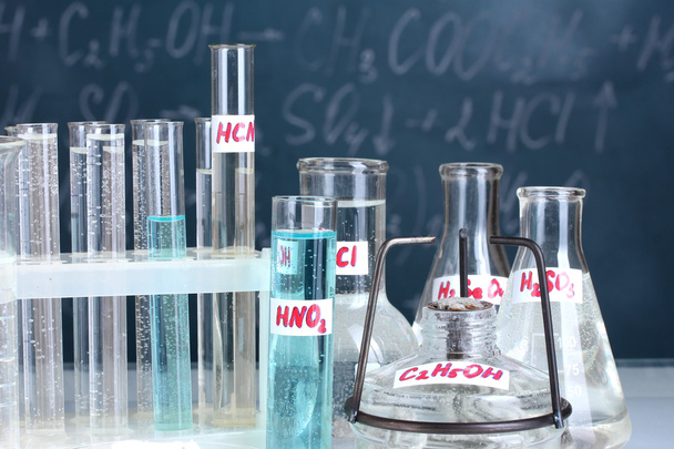 Пробирки с различными кислотами и другими химикатами на фоне доски
 - Фото, изображение