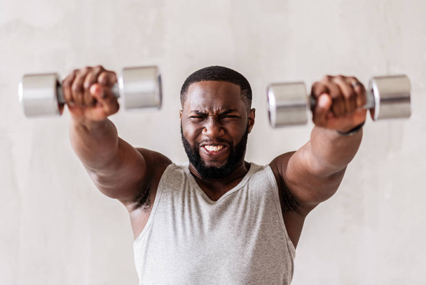 Angry young african sportsman performing exercises with hand weights - 写真・画像