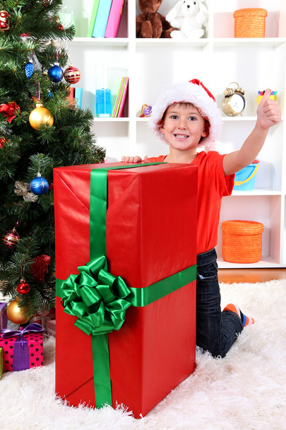 Child in Santa hat near Christmas tree with big gift - Foto, afbeelding