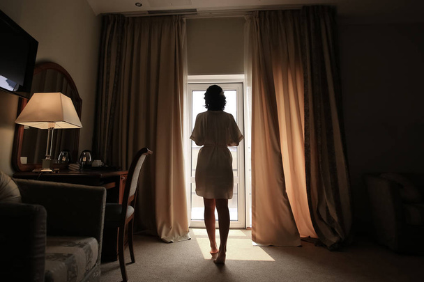 Beautiful young woman, silk bathrobe, standing by the window, natural light - Фото, изображение