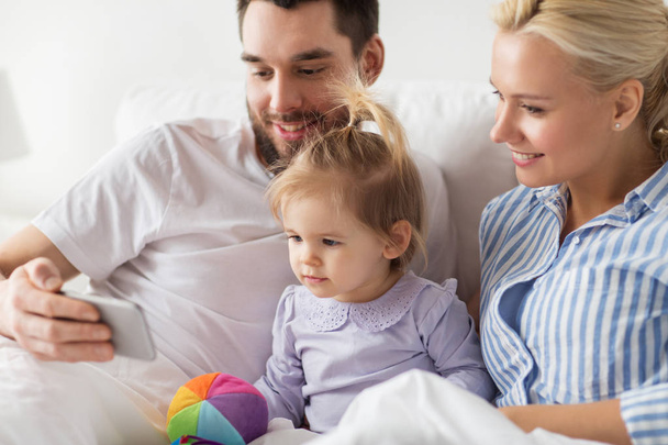 happy family with smartphone in bed at home - Foto, afbeelding