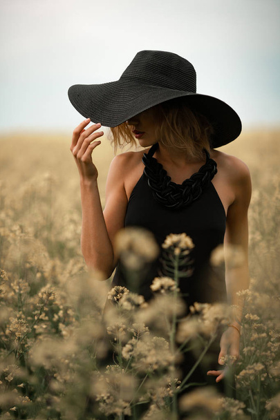 Siyah elbise ve şapka çiçekli çayır arasında genç kadın. - Fotoğraf, Görsel