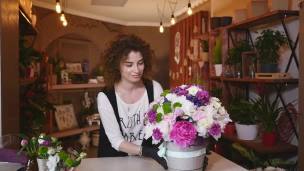 young woman working as florist in flower shop and looking at camera, smiling with bouquet on desk - Filmati, video