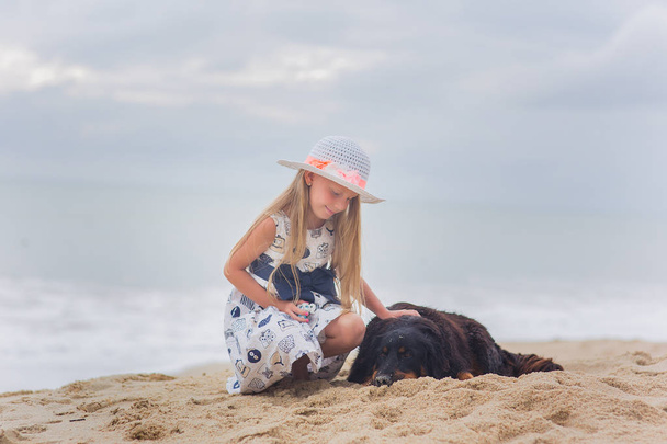 Cheerful pretty little girl in hat sitting and hugging her dog on the beach - Fotografie, Obrázek