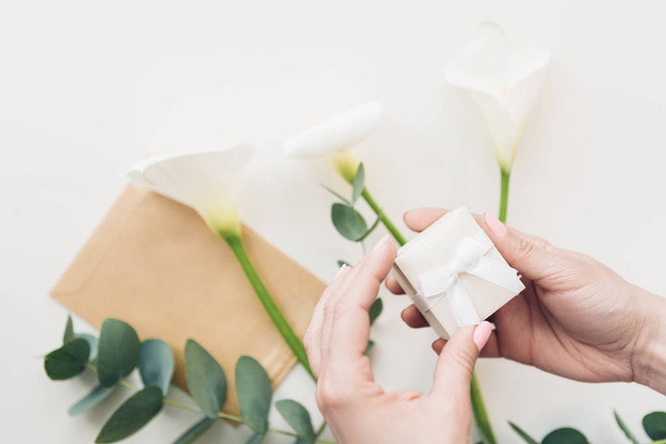 Female hands hold white gift box on background of flowers and phone. top view - Foto, afbeelding