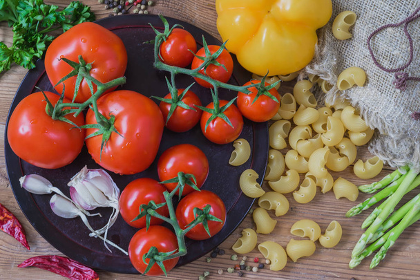 Ingredients for italian food - uncooked Italian pasta and vegetables, different varieties of tomatoes, garlic, asparagus, bell pepper on wooden background. Top view - Foto, Imagen