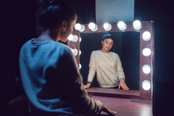 Attractive brunette in a gray sweatshirt looks in the mirror. - Фото, изображение