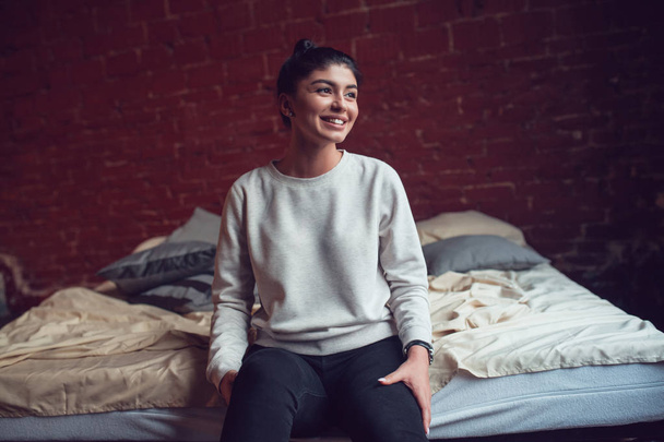 Attractive brunette woman in gray sweatshirt sits on bed. Mock-up. - Фото, изображение