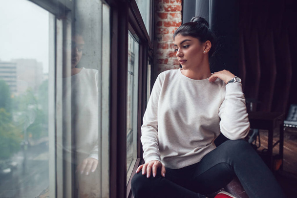 Attractive woman in gray sweatshirt sits on window sill and looks out the window. Mock-up. - Foto, imagen
