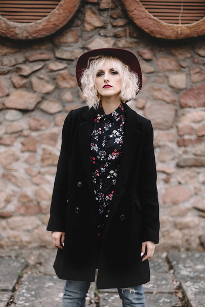 Beautiful cute blonde girl with curly hair in a hat and black coat near a stone wall - 写真・画像