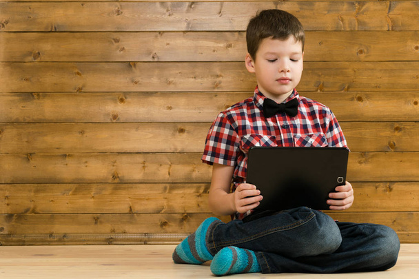 The boy sits on the wooden floor, and plays in the tablet - Фото, изображение