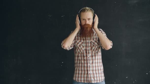 Portrait of bearded young man puts on headphones and dancing while listen to music on black background - Filmagem, Vídeo