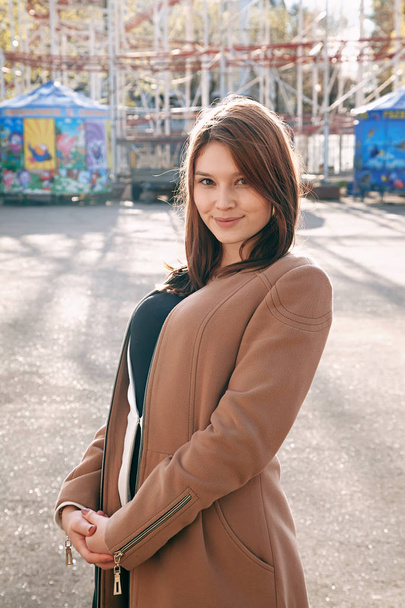Young girl posing in camera, smiling. City amusement park. In a brown coat of jeans - Фото, зображення