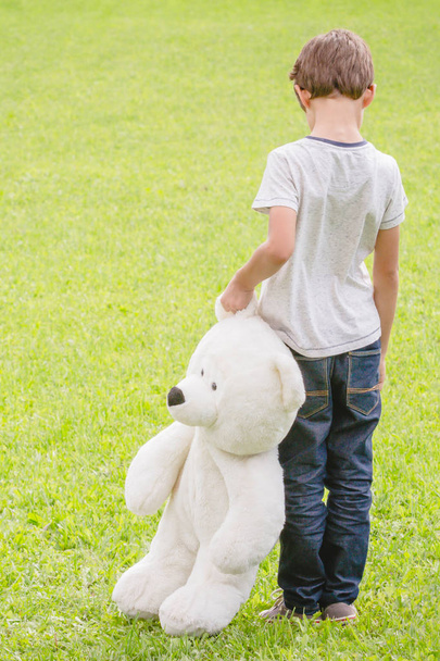 Sad lonely boy with teddy bear standing in the meadow. Child looking down. Back view. Sadness, fear, loneliness concept - Foto, imagen