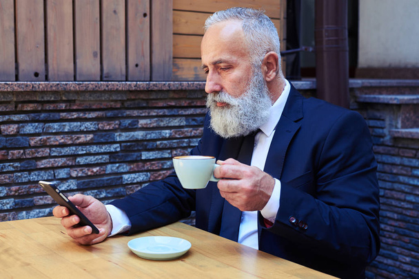 Przystojny stary człowiek pije kawę podczas surfowania w telefonie komórkowym - Zdjęcie, obraz