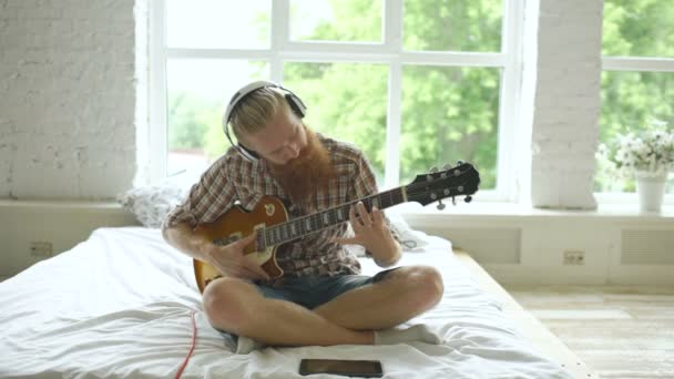Attractive bearded man in headphones sitting on bed learning to play guitar using tablet computer in modern bedroom - Filmagem, Vídeo