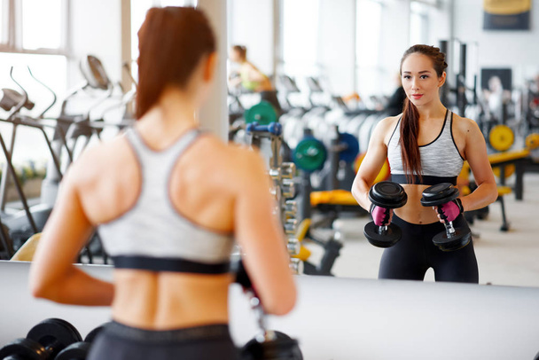 Concept of sport and healthy lifestyle. Strong young beautiful woman is engaged in a sports hall with dumbbells in front of a mirror - Photo, Image