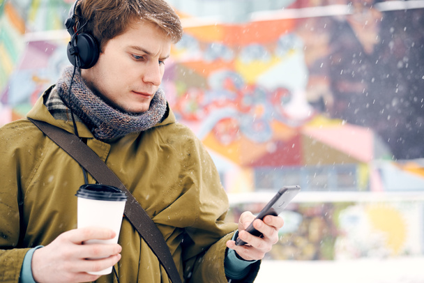 Man listening to music with headphones looking to the phone and drinking coffee in the winter on the street. Concept of technology and lifestyle - Photo, Image