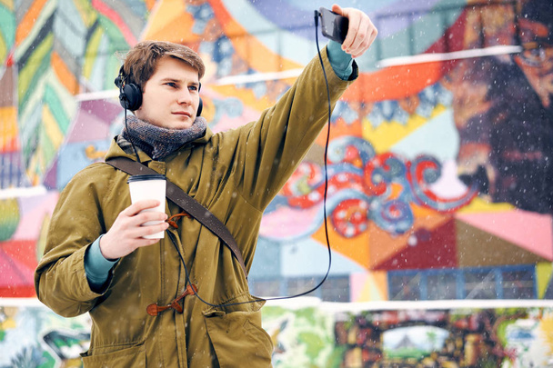 Мужчина слушает музыку в наушниках, фотографирует и пьет кофе зимой на улице. Концепция технологии и образа жизни
 - Фото, изображение