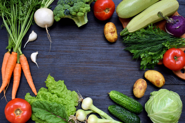 Σύνθεση σε σκούρο φόντο των οργανικών χορτοφαγικά προϊόντα: πράσινα φυλλώδη λαχανικά, καρότα, κολοκυθάκια, πατάτες, κρεμμύδια, σκόρδο, ντομάτες. Το Top view. Επίπεδη θέσει. - Φωτογραφία, εικόνα