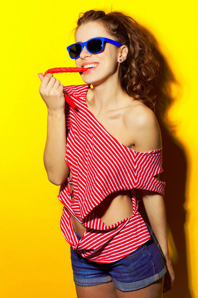 Menina sexy bonita em óculos de sol azuis e uma camiseta listrada vermelha rindo e se divertindo e comendo um doce em um fundo amarelo
 - Foto, Imagem