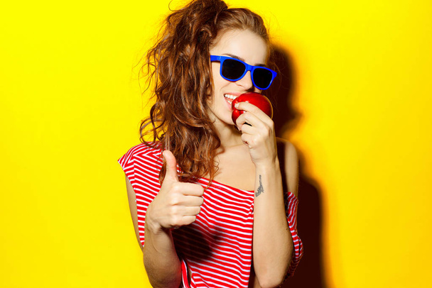 Beautiful young sexy girl in blue sunglasses and red striped T-shirt laughing and having fun and eating an apple on a yellow background - Фото, изображение