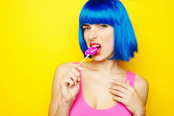 Mooie jonge sexy meisje in blauwe pruik en roze badpak met plezier en lolly snoep eten op gele achtergrond - Foto, afbeelding