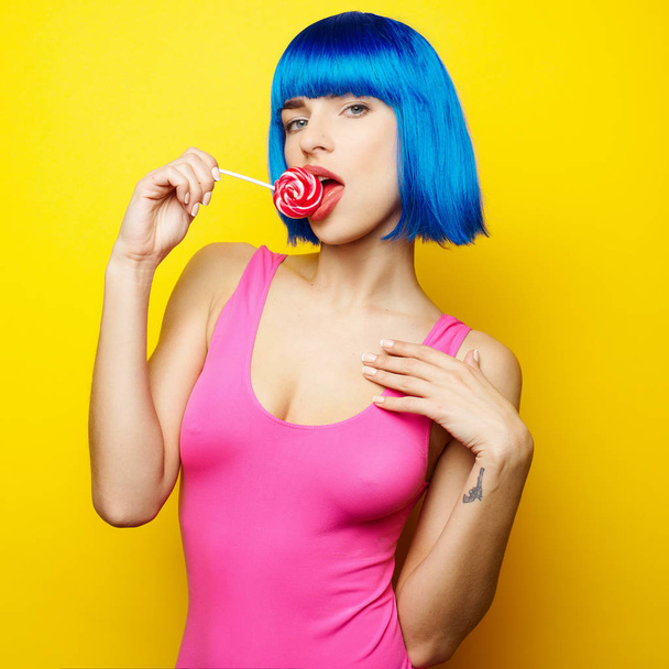 schöne junge sexy Mädchen in blauer Perücke und rosa Badeanzug Spaß haben und essen Lutscher Bonbons auf gelbem Hintergrund - Foto, Bild