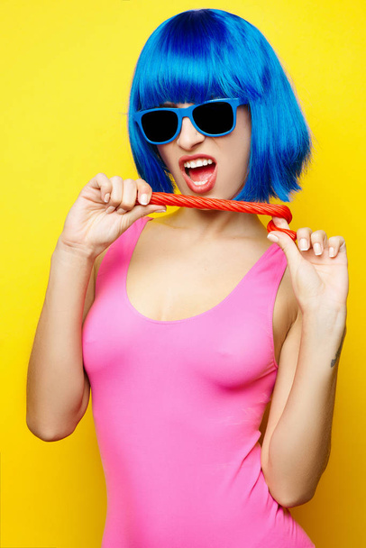 Beautiful young sexy girl in blue wig blue sunglasses and pink bathing suit having fun and eating lollipop candy on yellow background - Fotó, kép