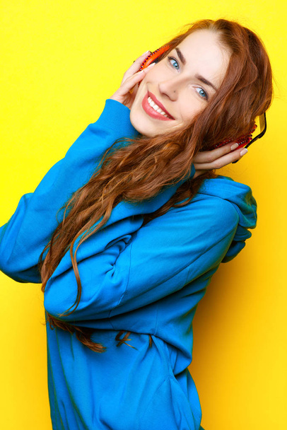 Beautiful young sexy girl DJ in a blue jacket listening music in red headphones on a yellow background and having fun - Photo, Image