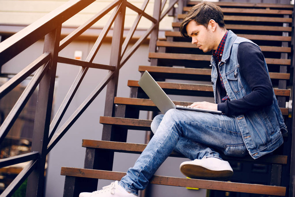 Genç adam merdivenlerde sokakta oturur ve bir dizüstü bilgisayar kullanarak. Yaşam tarzı ve teknoloji kavramı - Fotoğraf, Görsel