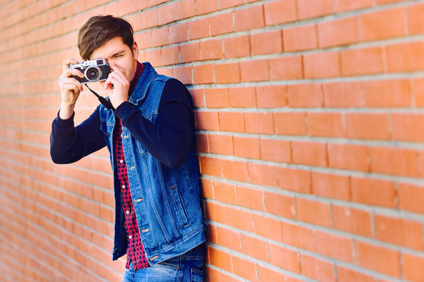 Young and handsome guy stands by a brick wall and takes pictures on a mirrorless camera - Foto, Imagen