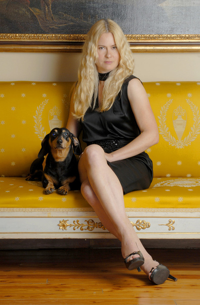 Young blonde woman with a little dog sitting on an elegant yellow sofa. - Фото, изображение