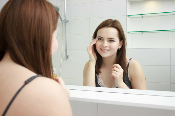 morning and people concept - young woman looking to mirror at home bathroom - Foto, Imagem