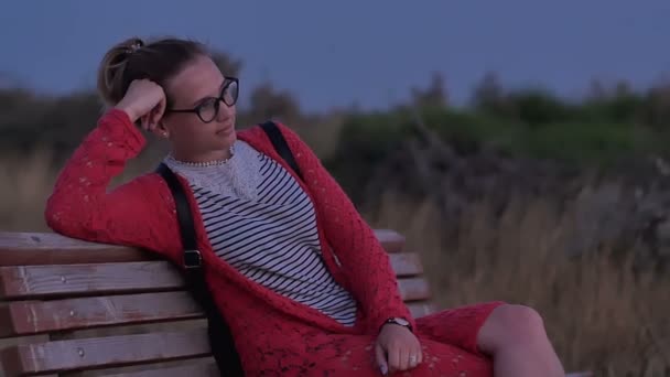 Девушка в очках сидит на скамейке и смотрит на вечернее море
 - Кадры, видео